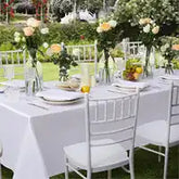 Elegant outdoor dining table set with white linens, silver chairs, and floral centerpieces.